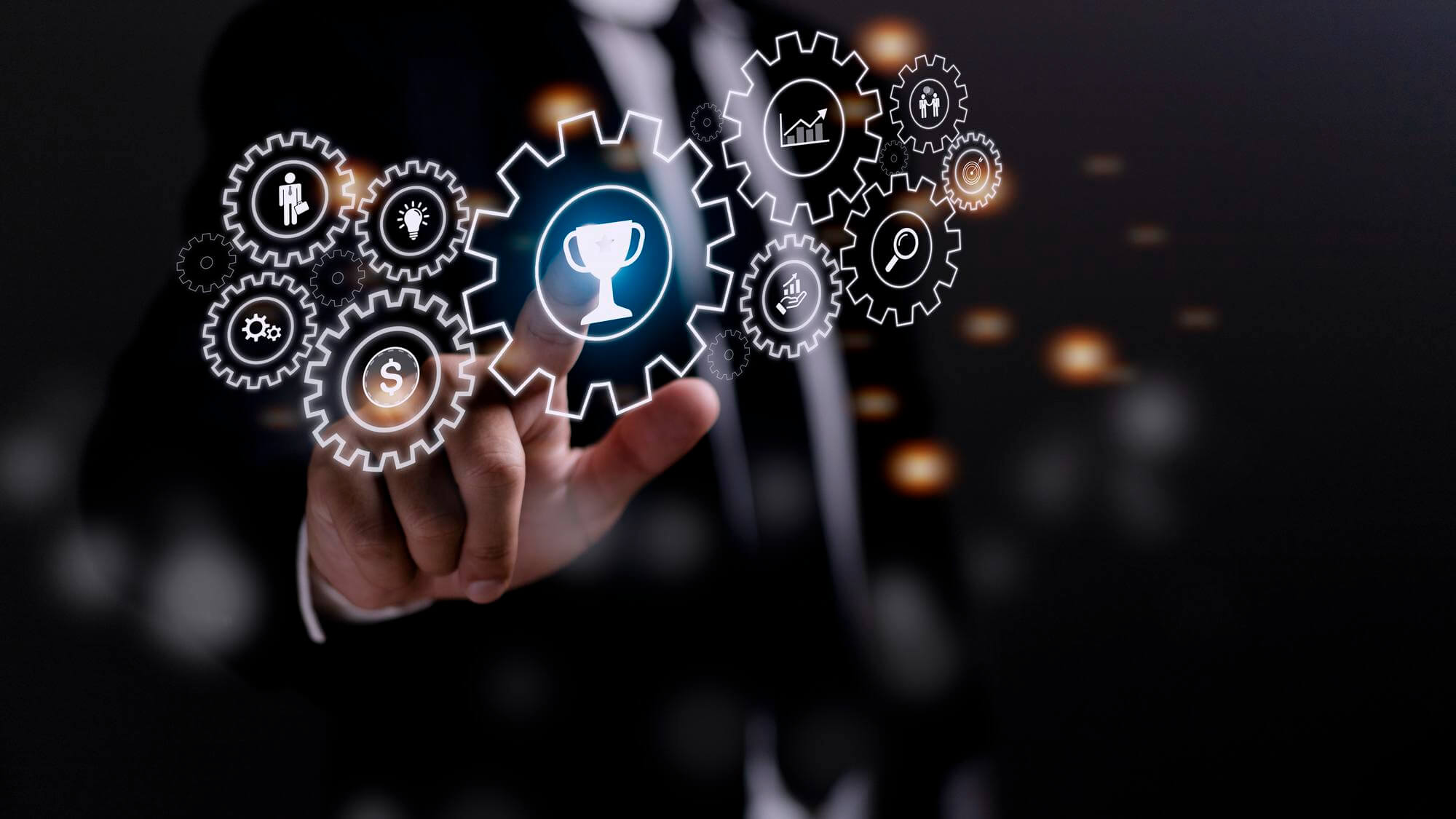 A man in a suit is touching a glowing gear with a trophy icon, surrounded by other gears with various business-related icons. The lighting is focused on the gears and the man's hand, with a dark background.
