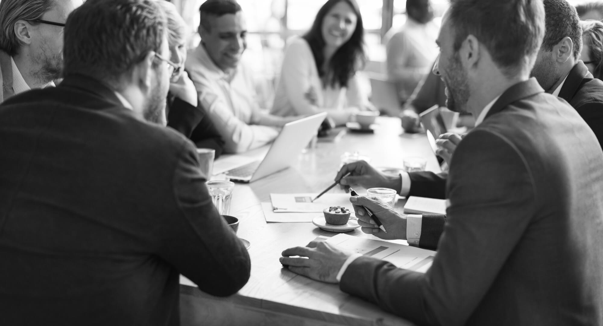 A black and white image shows a group of business people sitting around a table in a meeting, with a laptop, papers, and refreshments on the table.